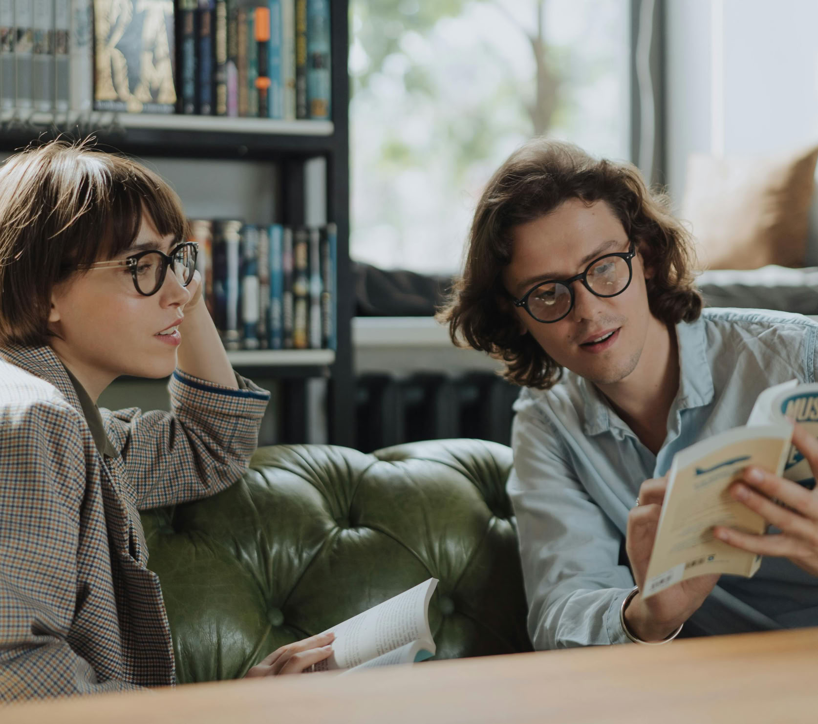 Two young adults in a library discussing books on a comfortable green sofa.